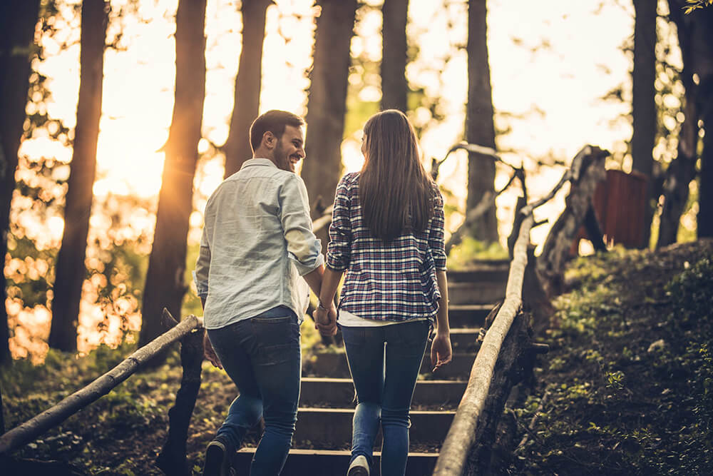 Couple hand in hand walking up a path outdoors together, smiling and looking at each other